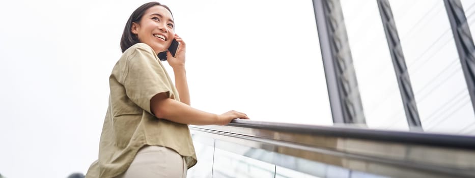 Happy asian woman on escalator, talking on mobile phone, meeting someone in city. Copy space