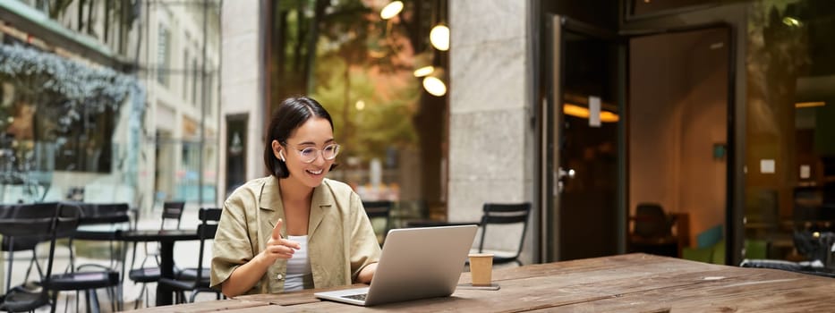 Young woman sitting on online meeting in outdoor cafe, talking to laptop camera, explaining something, drinking coffee.