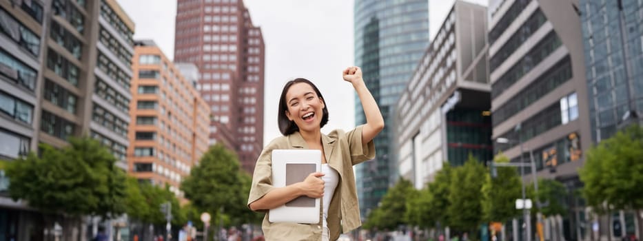 Portrait of happy asian woman stands with tablet near street road, cheering, raising hand up in triumph, celebrating.