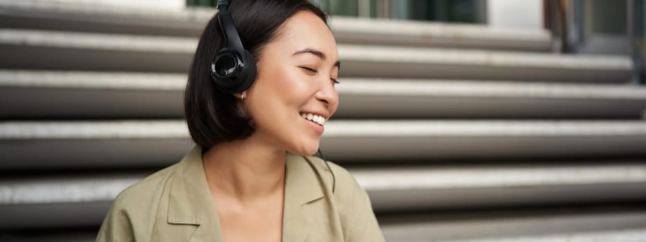 Young asian woman listening to music in headphones, sitting outside on street, smiling.