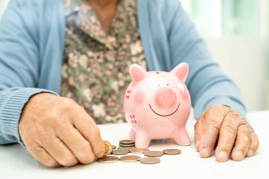 Asian elderly woman putting coin into pink piggy bank for saving money and insurance, poverty, financial problem in retirement.