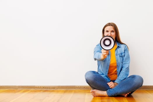 Young beautiful woman screaming on a megaphone sitting on a wooden floor in front of white wall.