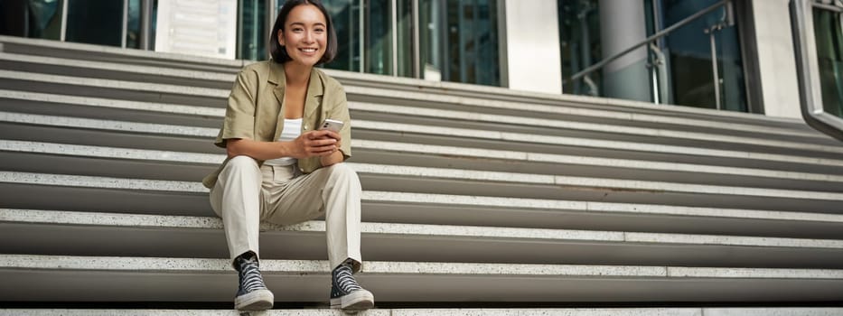 Portrait of smiling asian girl sits on stairs outdoors, sending message, using smartphone app, looking at mobile phone screen.