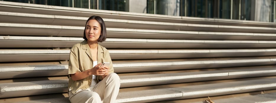 Beautiful asian girl sitting on stairs outside building, using mobile phone, looking at smartphone app and smiling.