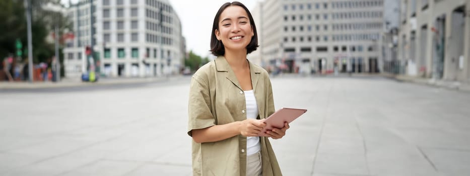 Young smiling asia woman with tablet, standing on street in daylight.