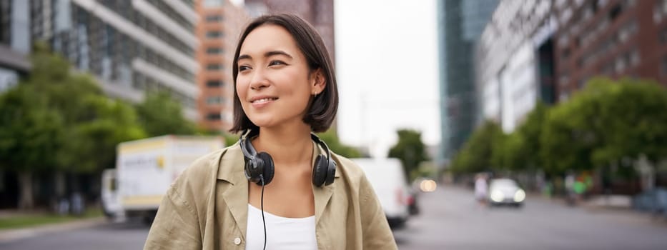 Portrait of young asian woman in headphones, posing in city, smiling and looking away, standing on street of city centre.