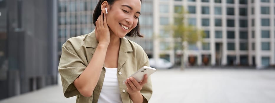 Smiling asian girl listens music in wireless headphones, looks at her phone, choosing music or podcast. Young woman calling someone, using headphones.