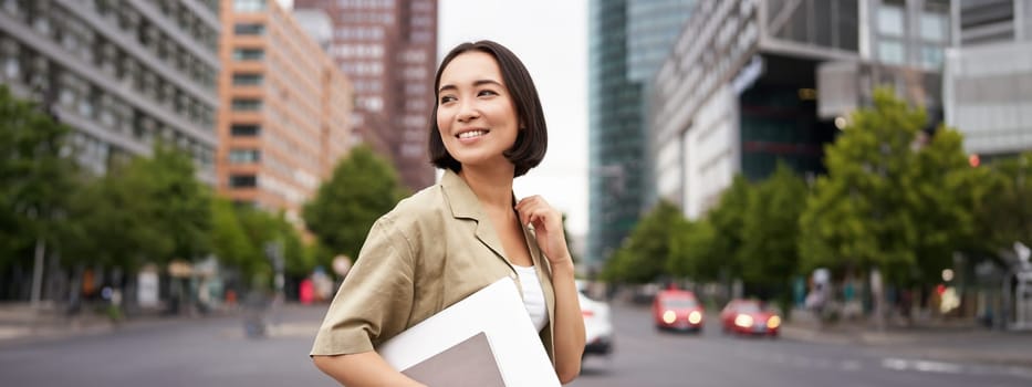 Outdoor shot of asian girl with laptop, going somewhere in city centre, walking on street, going to work.