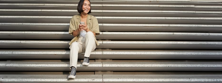 Portrait of smiling asian girl sits on stairs with her smartphone, browsing internet on mobile phone, resting outdoors in city centre.