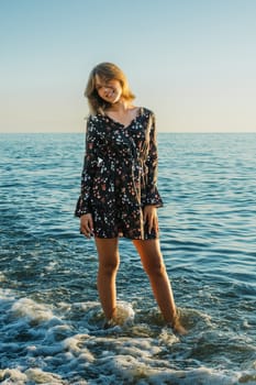 Teenage girl stands in a dress against the backdrop of the sea at sunset.