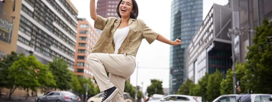 Portrait of dancing asian woman, raising hands up and feeling happy, posing in city, triumphing, feeling joy. Copy space