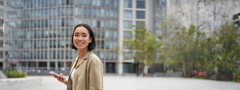 Vertical shot of asian girl walks with laptop on city street, smiles.