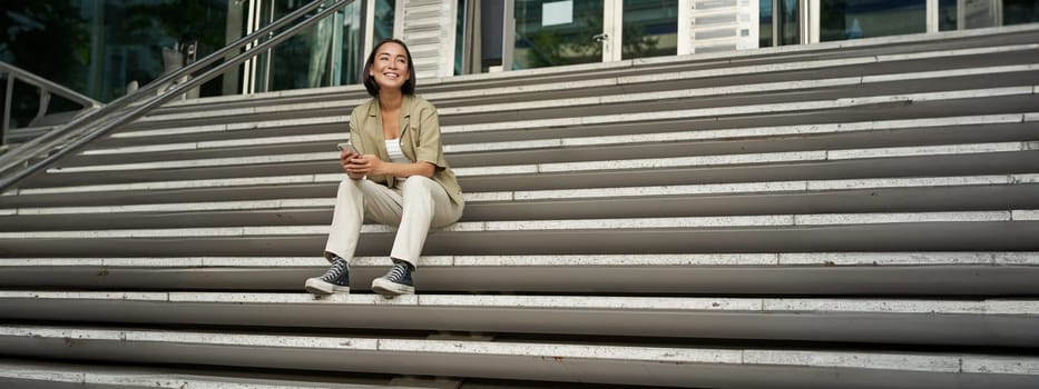 Portrait of smiling asian girl sits on stairs outdoors, sending message, using smartphone app, looking at mobile phone screen.