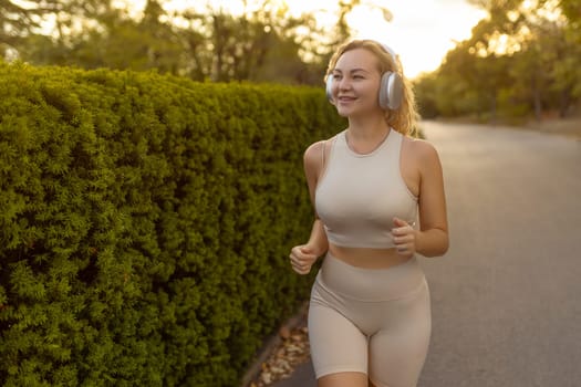 Smiling blonde woman wearing modern wireless headphones running through the park on a bright sunny summer day. A happy girl with a sweet smile runs and listens to good music. Lifestyle.