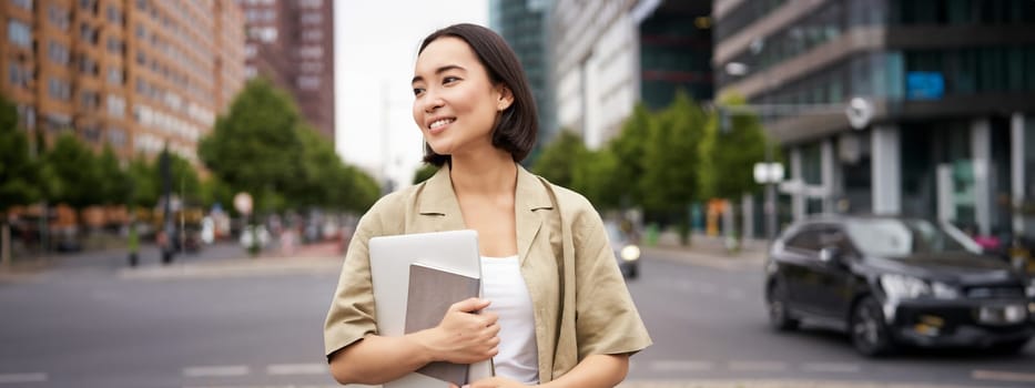Portrait of beautiful asian woman standing on street with laptop and notebook, going to university or work, commute route.