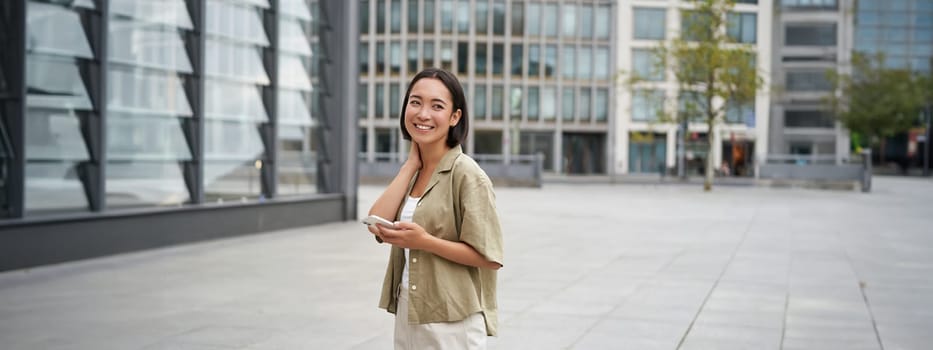 Portrait of asian woman walking in city. Street style shot of girl with smartphone, posing outdoors on street.