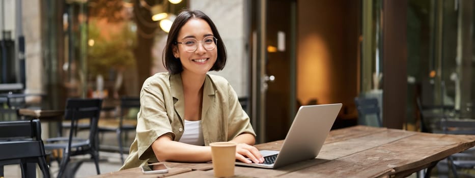 Portrait of smiling girl in glasses, sitting with laptop in outdoor cafe, drinking coffee and working remotely, studying online.