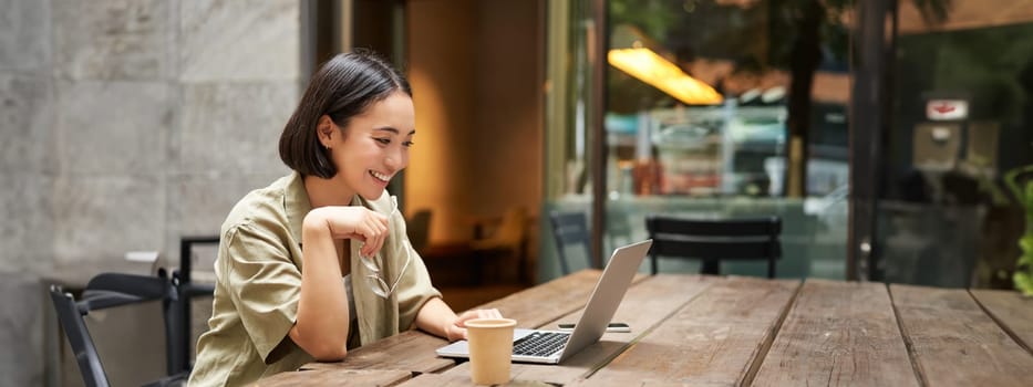 Portrait of asian woman looking at laptop, video chat, talking with someone via computer camera, sitting in cafe and drinking coffee, online meeting.