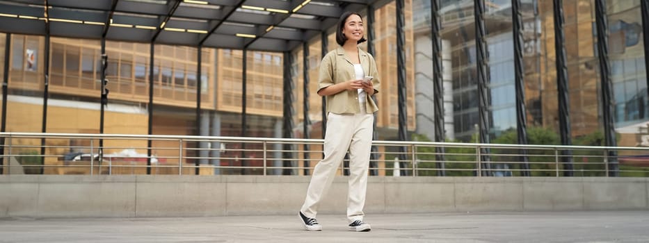Portrait of young asian woman, student with smartphone, standing on street near glass building, waiting for someone.