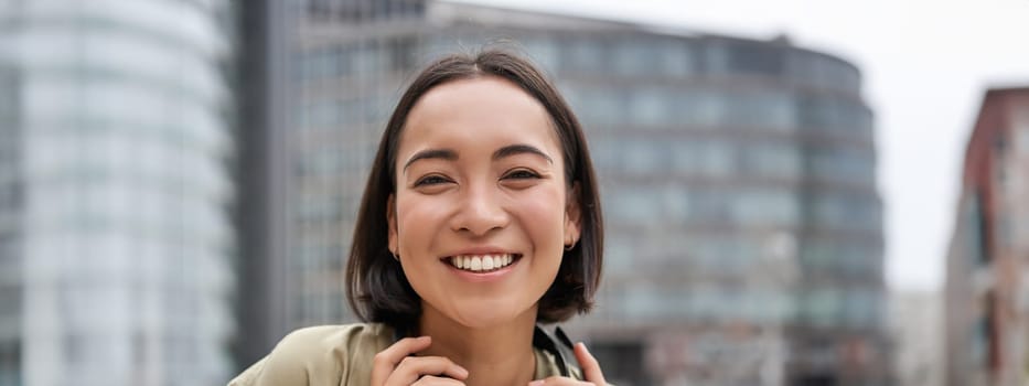 Vertical shot of beautiful asian woman posing with headphones around neck, smiling and laughing, standing on street in daylight.