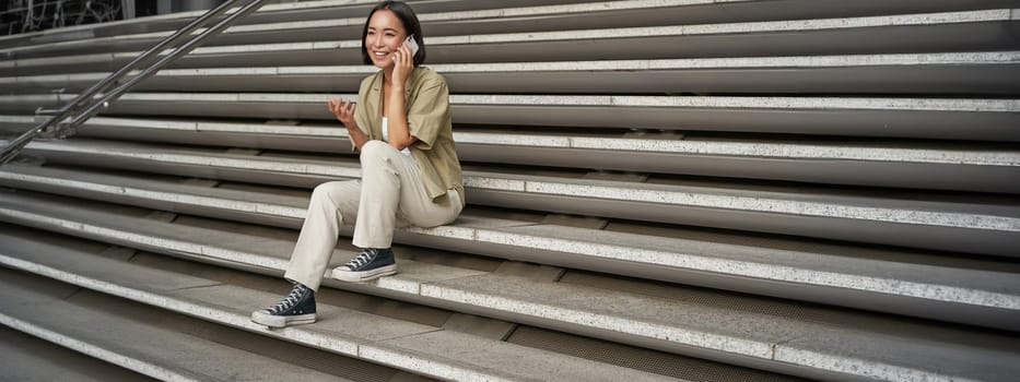 Smiling asian girl sits on stairs of building and talks on mobile phone, relaxing during telephone conversation.