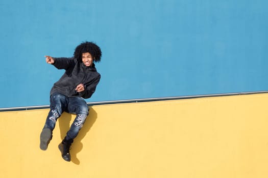 funny african american teenager smiling happy pointing to the side while sitting on a yellow wall in front of blue background, concept of adolescence and young lifestyle, copy space for text