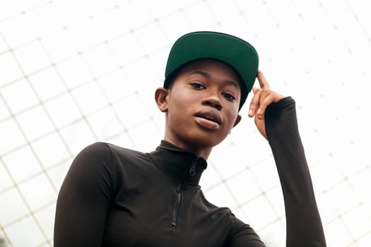 portrait of a young tender african american woman in casual wear touching her cap with finger with a blurred fence in the background