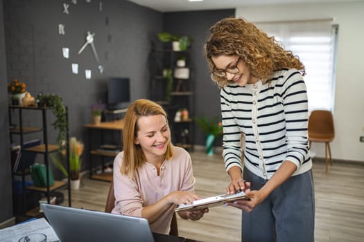 Two female colleagues are working from home, one is sitting at a desk using a laptop, while the other is standing next to her with a document in hand, working together on a business report.