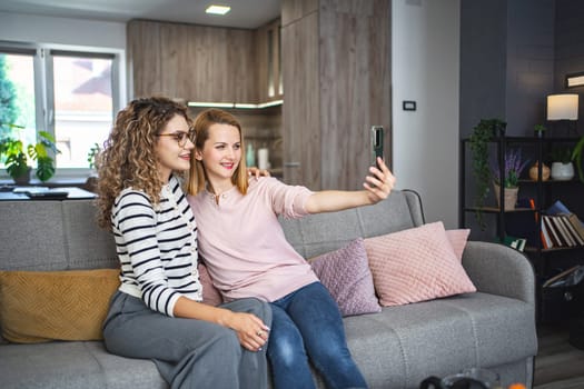 Two women are seated on a couch, holding a smartphone at arms length to take a selfie together. They are smiling while capturing the moment on the screen.