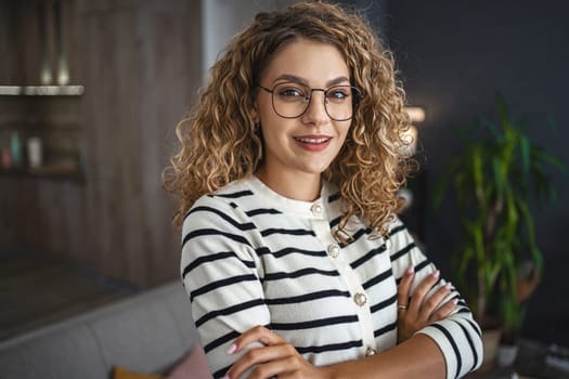 A woman wearing glasses is standing in front of a couch, looking at the camera. The couch is in the background, creating a homey atmosphere.