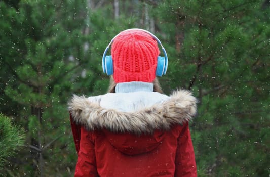 Back view modern woman enjoying listening to music with headphones in hat, jacket in winter forest against Christmas tree background with snow
