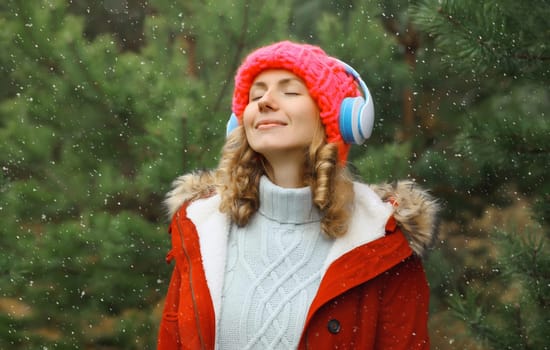 Portrait of happy modern young woman enjoying listening to music with headphones in hat, jacket in winter forest against Christmas tree background with snow
