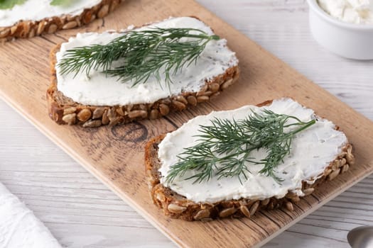 Two slices of rustic rye bread are generously spread with creamy topping, garnished with fresh dill, placed on a wooden board in a well-lit kitchen.