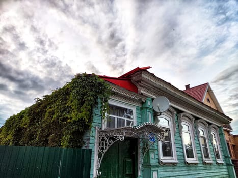 A beautifully preserved wooden house covered in greenery features intricate architectural details against a dramatic cloudy sky.