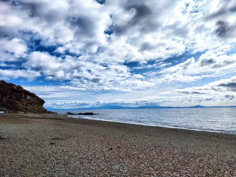 A peaceful coastline with pebbles and gentle waves under an expansive cloudy sky during sunset creates a serene atmosphere.