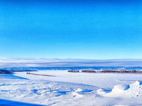 A sprawling expanse of snow blankets the ground, with gentle hills and distant mountains framed against a vibrant blue sky in winter.