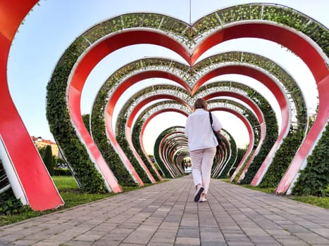 A walker strolls beneath decorative heart-shaped arches covered in greenery, enjoying a peaceful moment in the park during evening hours.