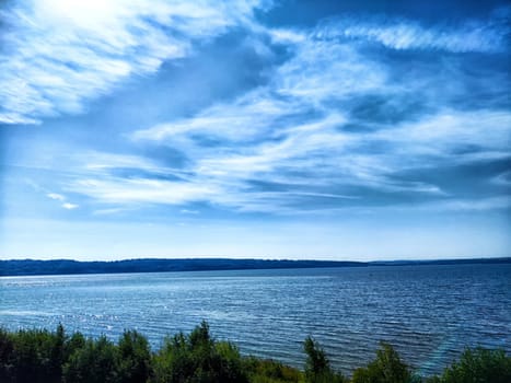 Blue skies stretch over a calm lake, reflecting soft clouds while trees line the tranquil shore on a sunny day.