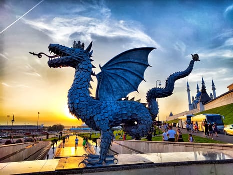 Kazan, Russia - July 04, 2024: The serpent Zilant statue stands majestically, backlit by stunning sunset over the Kazan riverfront, captivating onlookers nearby