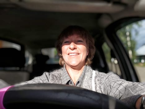 A middle aged woman enjoys her drive, smiling happily as she navigates the road on a bright, sunny day.
