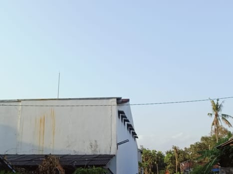 A rustic building exterior with visible rust stains is set against a clear blue sky. Coconut tree and greenery surround the structure, evoking a tropical, serene atmosphere.