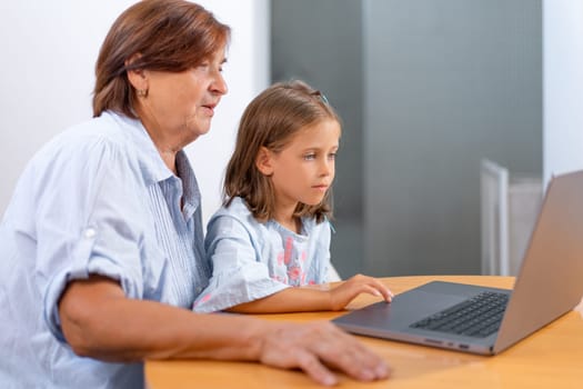 Focused granddaughter and grandmother surfing on laptop together at home. Senior woman and girl are learning from the computer during leisure time