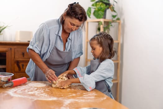 Cute girl kneading pastry dough on table while baking with grandmother at home. Little female child is learning baking from elderly woman, baking concept