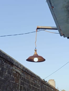 A rustic outdoor light fixture suspended from a building against a clear blue sky. The weathered lamp exudes a sense of simplicity and rustic charm, casting a warm, inviting glow.