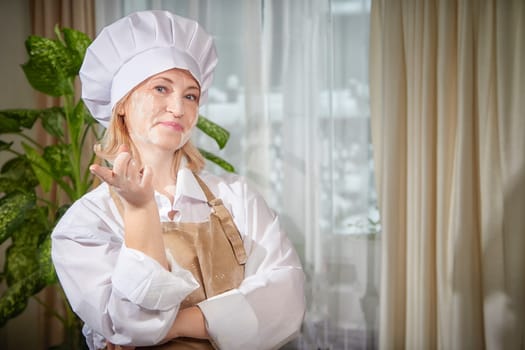 An elderly woman dressed as a cook is playfully posing in a kitchen, surrounded by flour, showcasing a joyful moment.