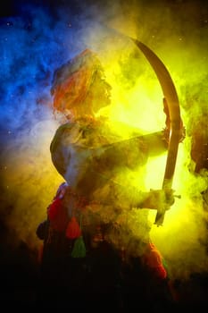 A woman in a colorful headscarf and jewelry poses With a saber in front of wall of colorful tassels and smoke around