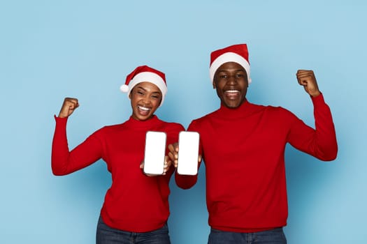 A joyful black couple, wearing matching red sweaters and Santa hats, proudly holds up their smartphones while smiling enthusiastically against a light blue background.