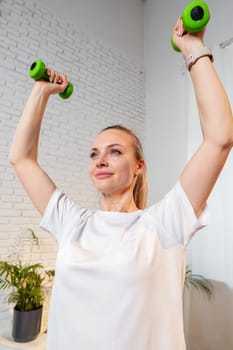 A woman exercises with green dumbbells at home in a bright room filled with light and plants