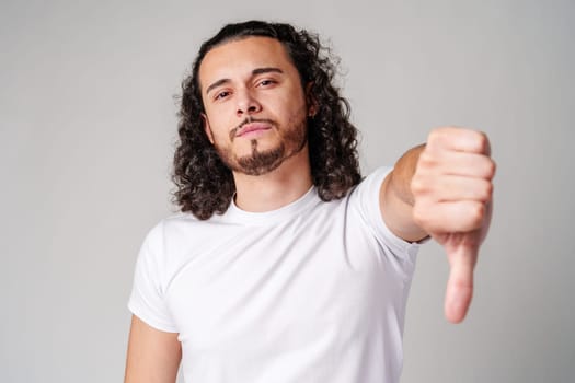 A young man with curly hair gestures disapproval by giving thumbs down in a neutral studio setting close up