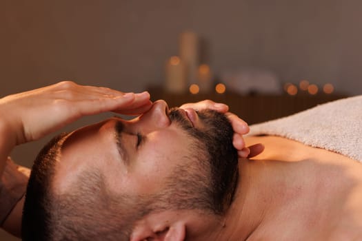 Close-up of female massage therapist's hands massaging man's forehead on dark background, effective facial massage for wrinkles, rejuvenation